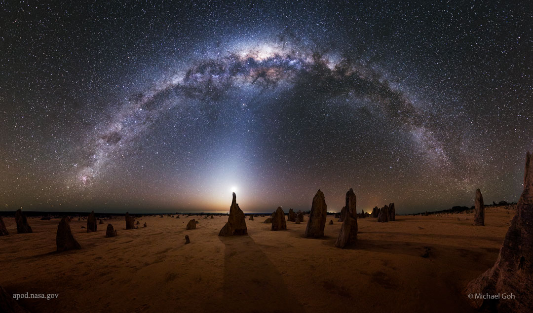 A flat landscape is shown at night that appears mostly  brown. Numerous unusual rock spires are seen rising from the   group. Above, a full star field is seen with the arch of our   Milky Way Galaxy curving from left to right.   Please see the explanation for more detailed information.