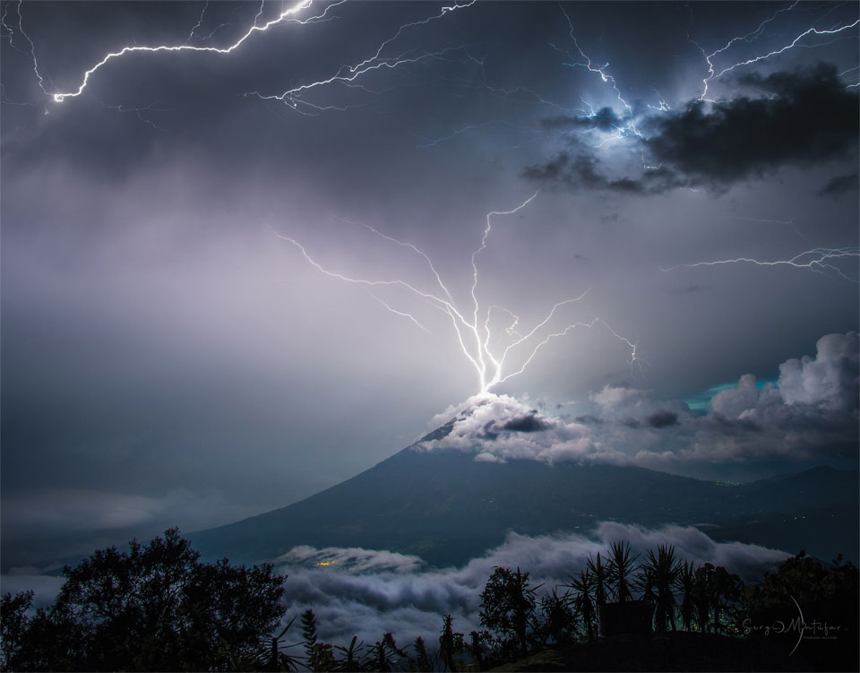 A volcano is shown with its peak in the midst of 
purple clouds. Lightning appears to come out of the 
top of the volcano in multiple paths into the upper sky.
Please see the explanation for more detailed information.