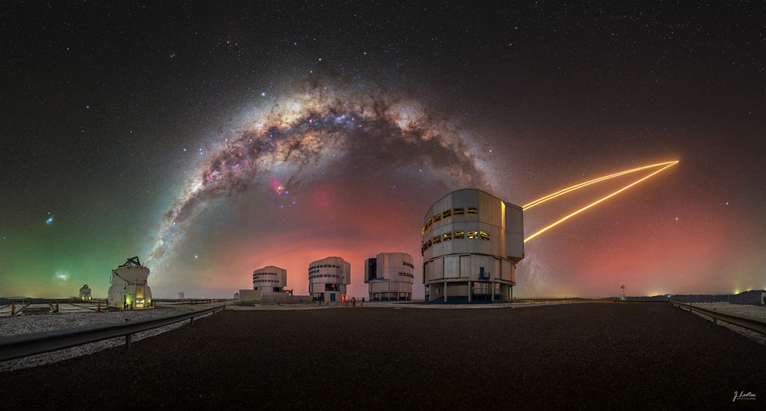 A colorful sky appears above several large telescopes.
Prominent in the sky is the band of the Milky Way Galaxy which
arches across the frame. Orange laser beams connect one
of the telescopes to the sky on the image right. 
Please see the explanation for more detailed information.