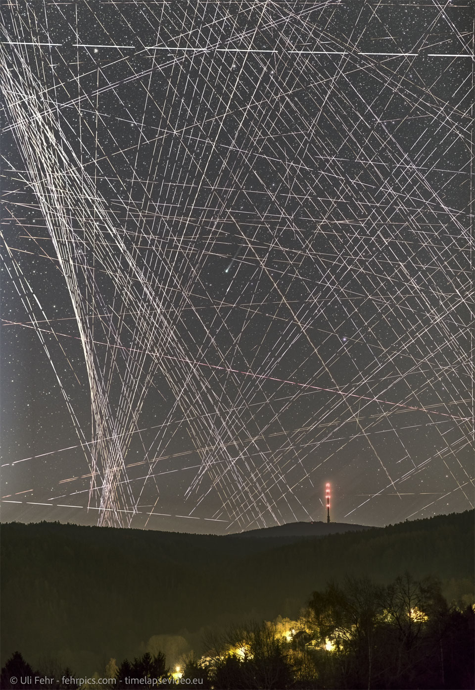 A night sky filled with lines and dashed-lines 
appears above a hilly landscape featuring a distant
lit-up tower. Near the center of the image is a comet
shown by its small green coma and ion tail to the upper 
right.
Please see the explanation for more detailed information.
